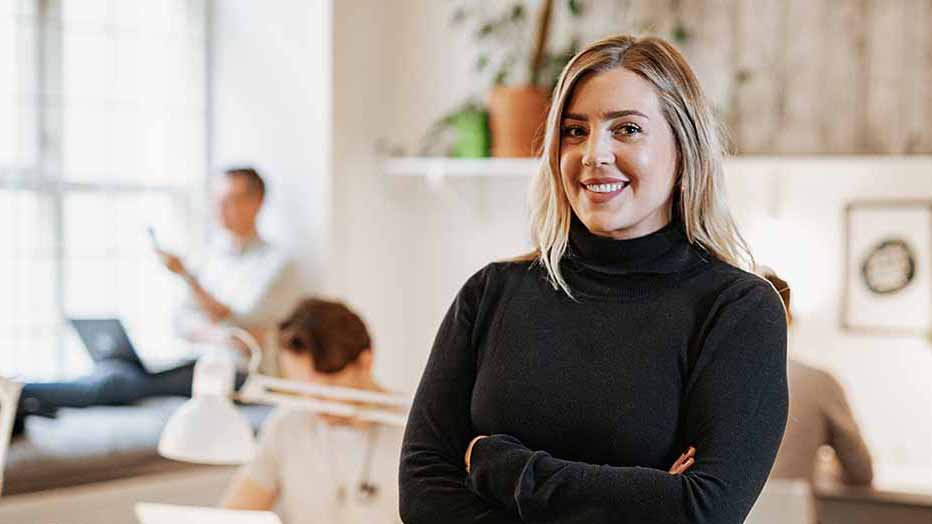 Young woman represents a team development coach. She smiles and stands with her arms crossed in an office landscape.